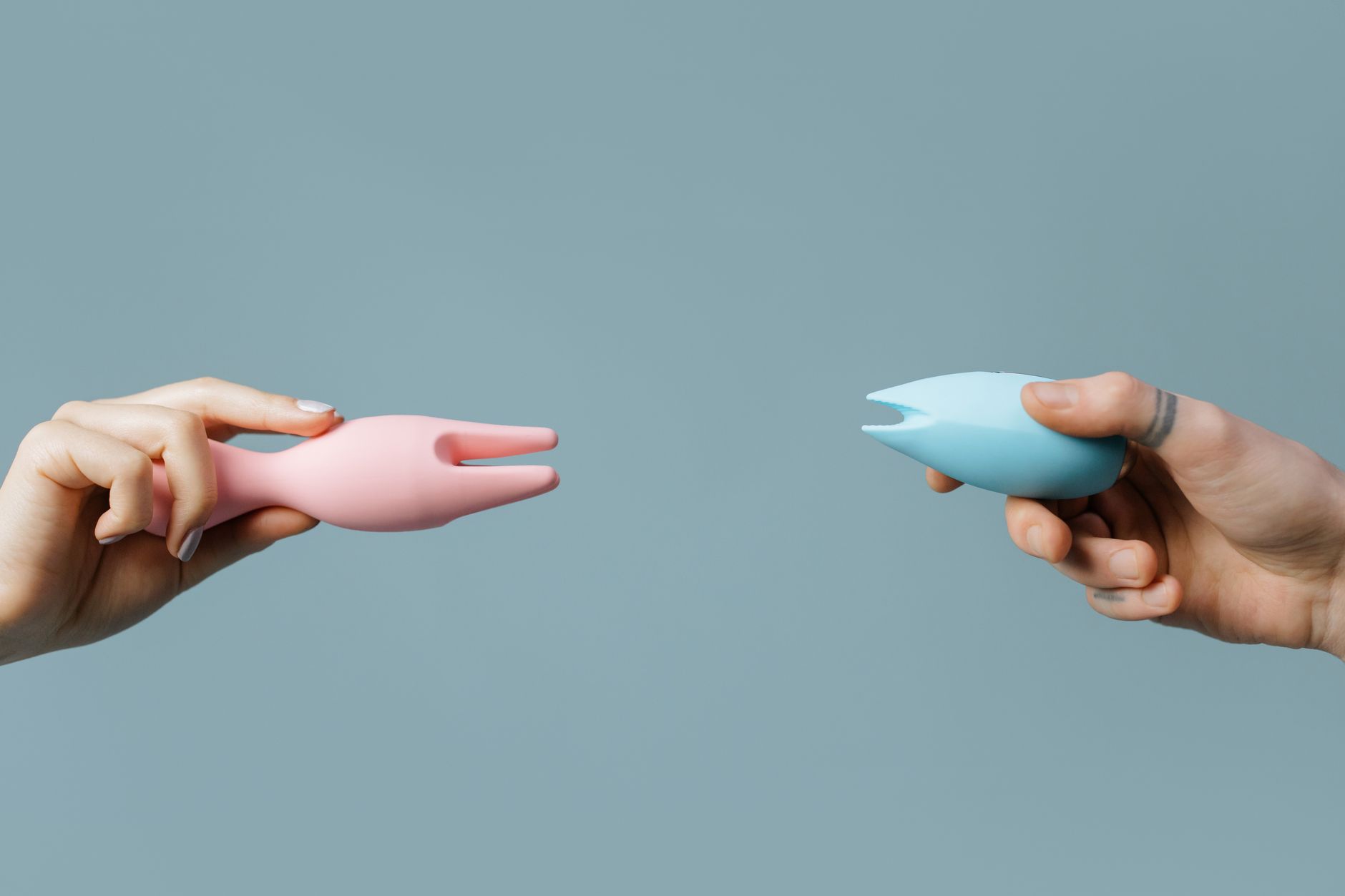 Hands holding pink and blue silicone vibrators against a pastel background